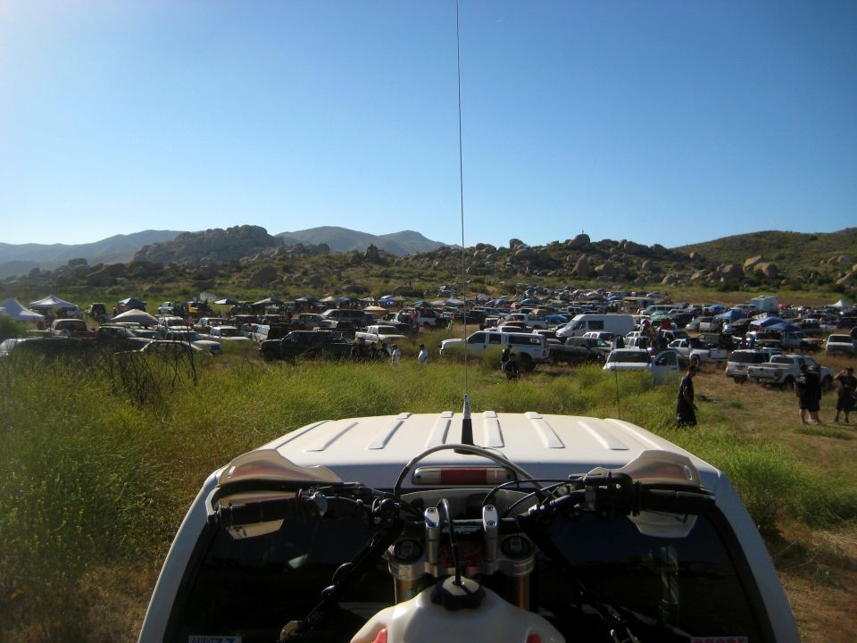 View from inside a vehicle looking out at a large parking area filled with many cars and trucks in a dry, hilly landscape