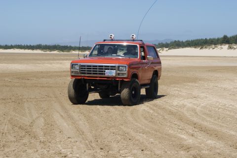 1983 Ford Bronco - Orange Ford Bronco with lifted suspension driving on a sandy beach under a clear blue sky.