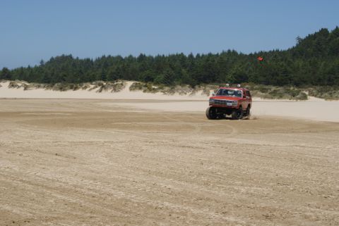 1983 Ford Bronco - A red SUV kicks up sand as it drives across a beach, with a line of trees and dunes behind it.