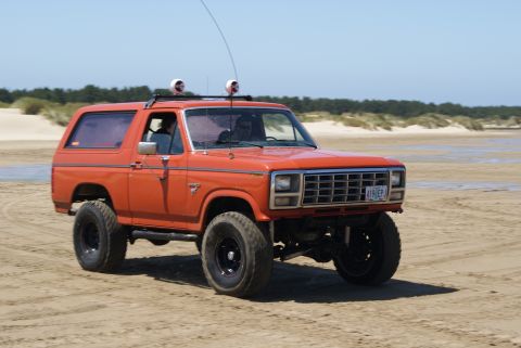 1983 Ford Bronco - This orange Ford Bronco is captured in motion on a sandy beach under a clear blue sky.