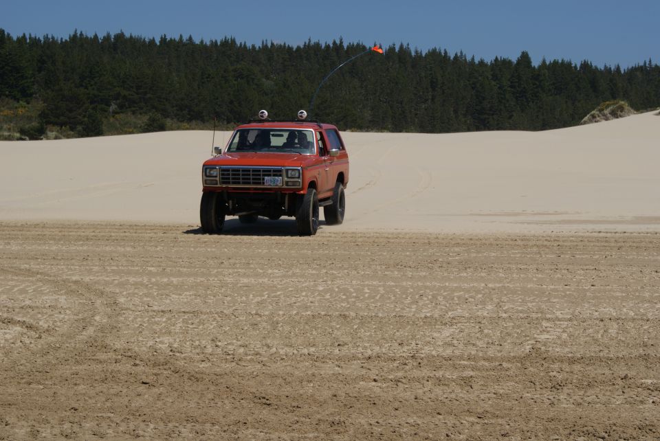 Orange 1983 Ford Bronco driving on sand dunes with an orange flag on an antenna
