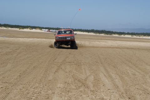 1983 Ford Bronco - An orange SUV kicks up sand as it drives along the beach near dunes and trees.