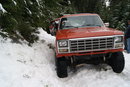 1983 Ford Bronco - This orange pickup truck is navigating a snowy, wooded area with a second vehicle visible behind it.