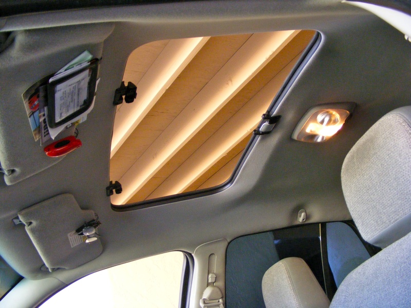 Car interior ceiling with open sunroof showing wood beams above, gray headliner, visors and lit dome light