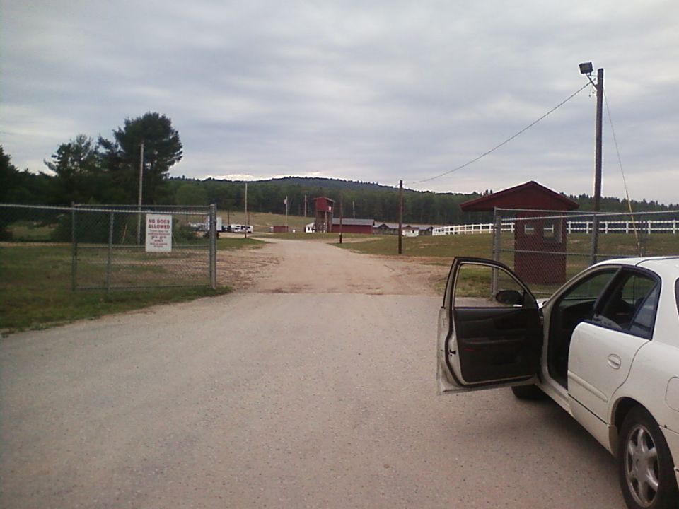 White sedan parked on dirt road, driver's side doors open. Buildings and trees in background