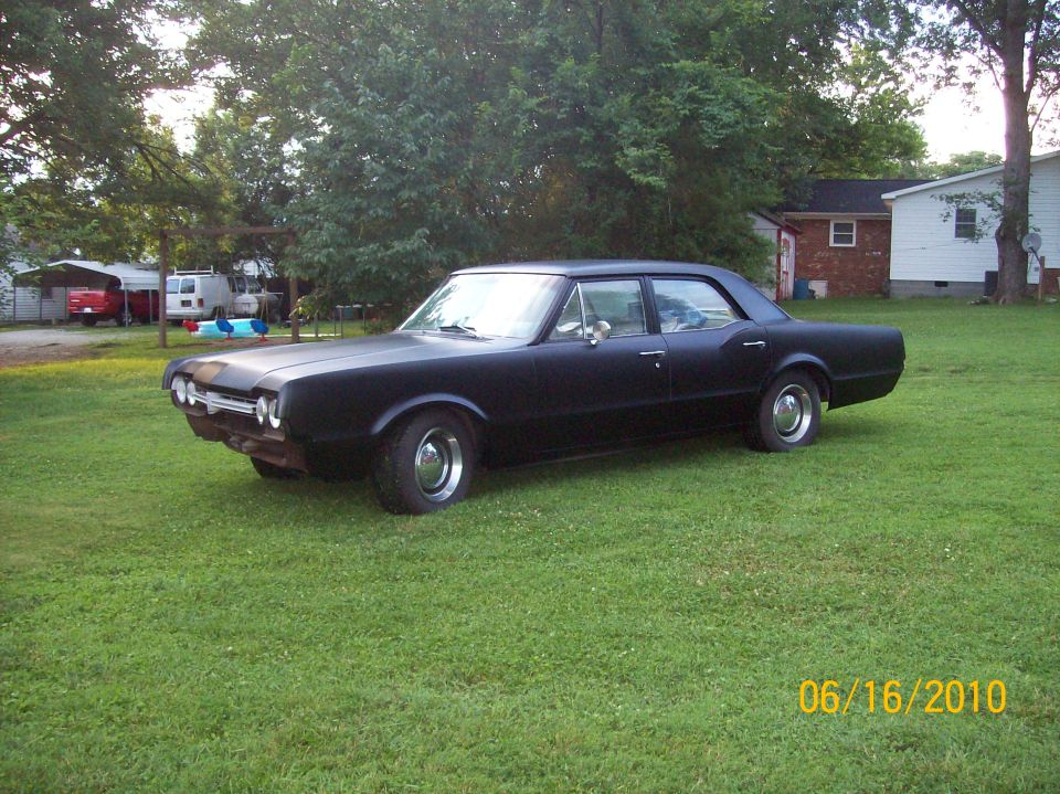 Flat-black 1960s Oldsmobile Cutlass four-door sedan parked on a grass lawn