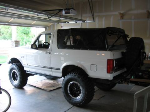 1995 Ford Bronco - A white two-door Ford Bronco with a black soft top parked inside a garage, shown from the rear three-quarter driver's side. It has a suspen…