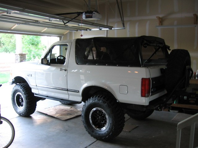 White Ford Bronco with black soft top, lifted on off-road tires, parked in a garage.