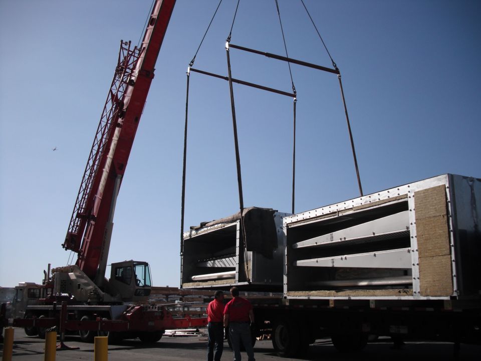 Crane lifting large metal boxes off a flatbed truck. Two workers in red shirts stand nearby