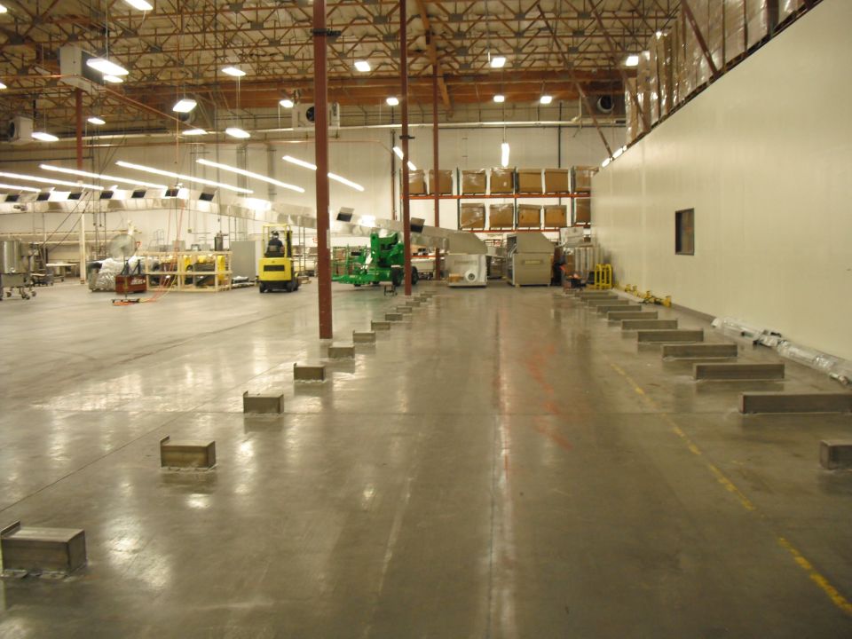 Warehouse interior with concrete floor, high ceiling, industrial lighting, and various machinery. Forklift visible