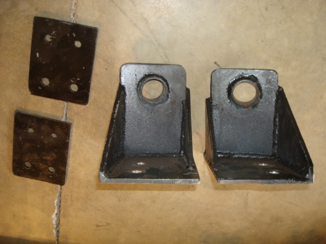 Four dark metal brackets on a light brown surface. Two are L-shaped with a circular hole, two are flat plates with four holes
