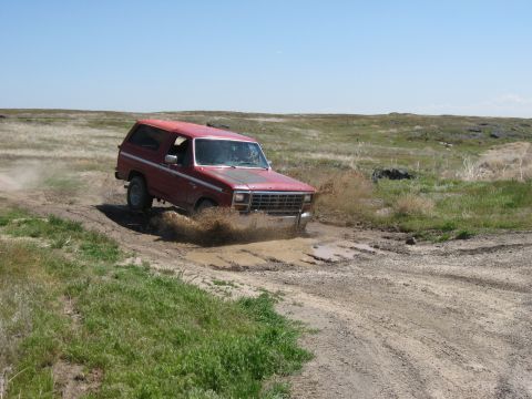1981 Ford Bronco - A red SUV navigates a muddy trail under a clear blue sky.
