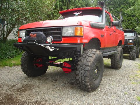 1989 Ford Bronco - This heavily modified red SUV features a custom front bumper, snorkel, and oversized tires, with another vehicle visible in the background.