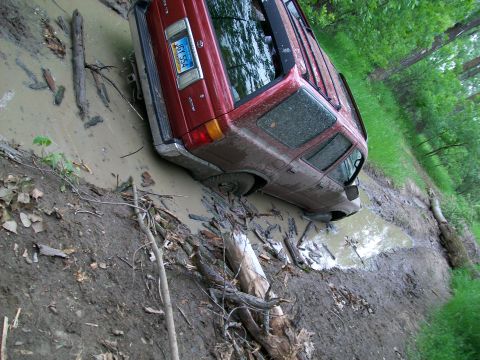 1988 Ford F-150 - A red SUV is stuck in a muddy ditch with trees and logs surrounding it.