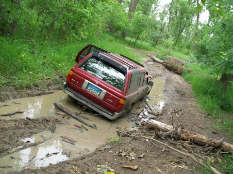 1988 Ford F-150 - A red SUV is stuck in a muddy rut on a dirt path surrounded by trees and vegetation.