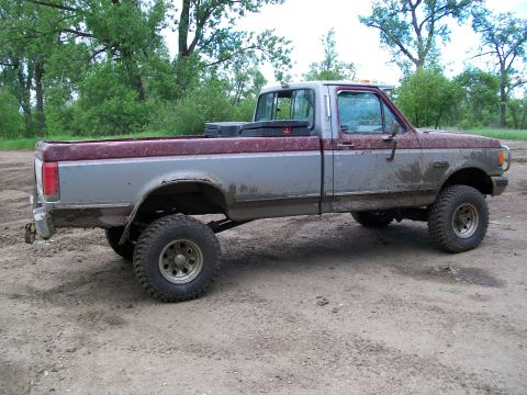 1988 Ford F-150 - This lifted Ford F-150 pickup truck is shown from the side, parked on a dirt surface with trees in the background.