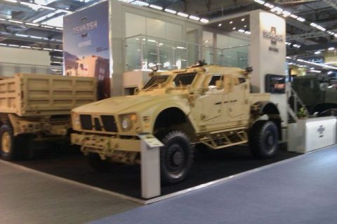 1991 Ford Crown Victoria - An Oshkosh M-ATV armored vehicle is displayed next to a dump trailer at an indoor exhibition.