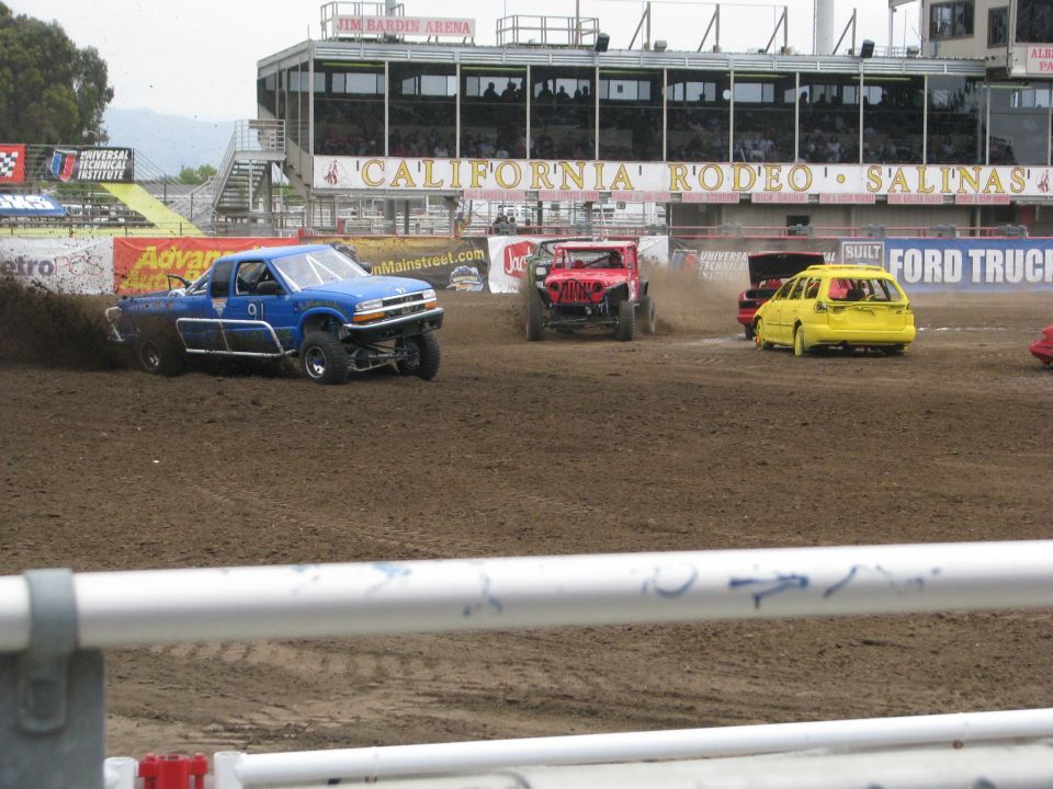 Blue pickup truck kicking up dirt on a race track, with other vehicles visible