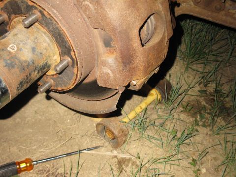 1978 Ford Bronco - Close-up view of a heavily rusted brake caliper and rotor assembly, with a yellow rod lying on the grass nearby.