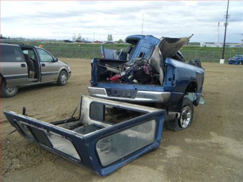 2005 Dodge 3500 - A severely damaged blue pickup truck sits with its camper shell removed, revealing a load of debris in the truck bed.