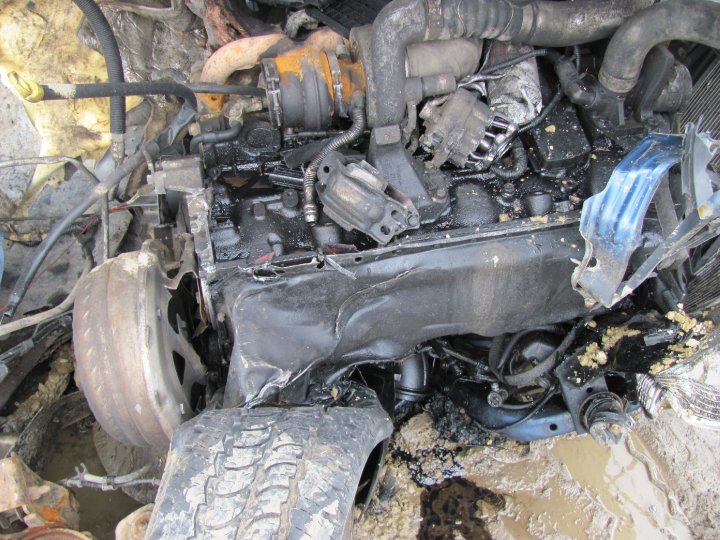 Close-up of a damaged engine compartment with a tire and wheel visible. The engine is covered in oil and grime