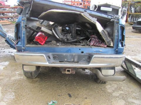 2005 Dodge 3500 - Rear view of a damaged blue pickup truck in a junkyard, filled with various parts and debris.