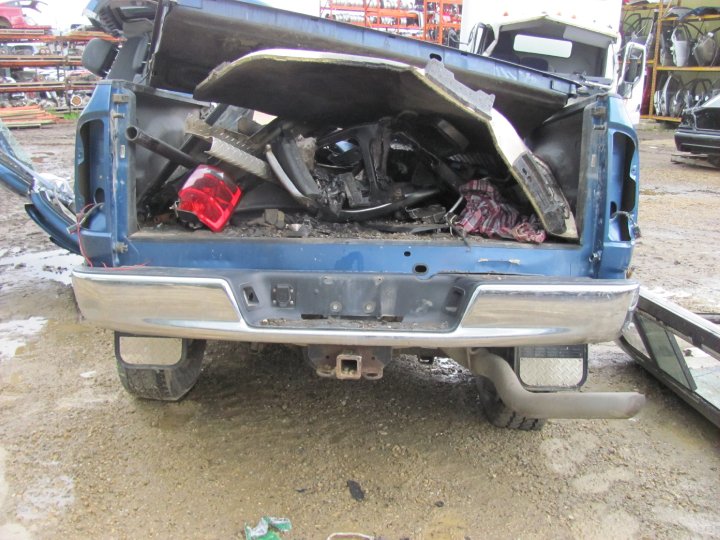 Blue pickup truck rear bumper and bed filled with debris. Chrome bumper, trailer hitch, and red taillight visible