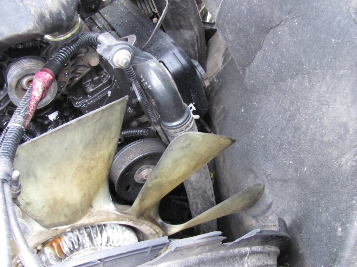 Engine fan blades and pulleys with black hoses and wiring. A grey textured surface is on the right