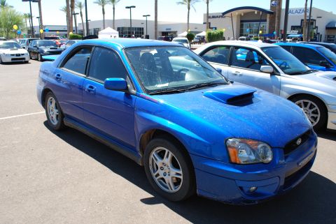 2005 Subaru Impreza - A blue 2005 Subaru Impreza WRX is parked in front of a Subaru dealership, alongside other vehicles.