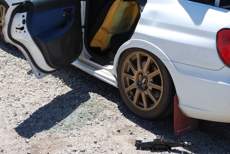 White 2005 Subaru Impreza side view with driver door open, showing blue interior trim and yellow foam padding. Gold BBS wheels and red mud flaps are visible