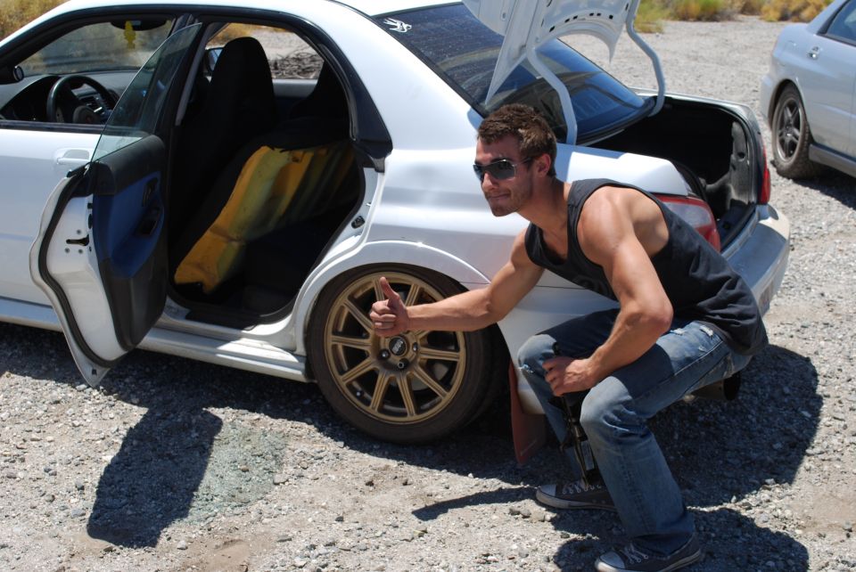 Man crouching with a jack beside a white Subaru WRX STi with gold wheels on gravel