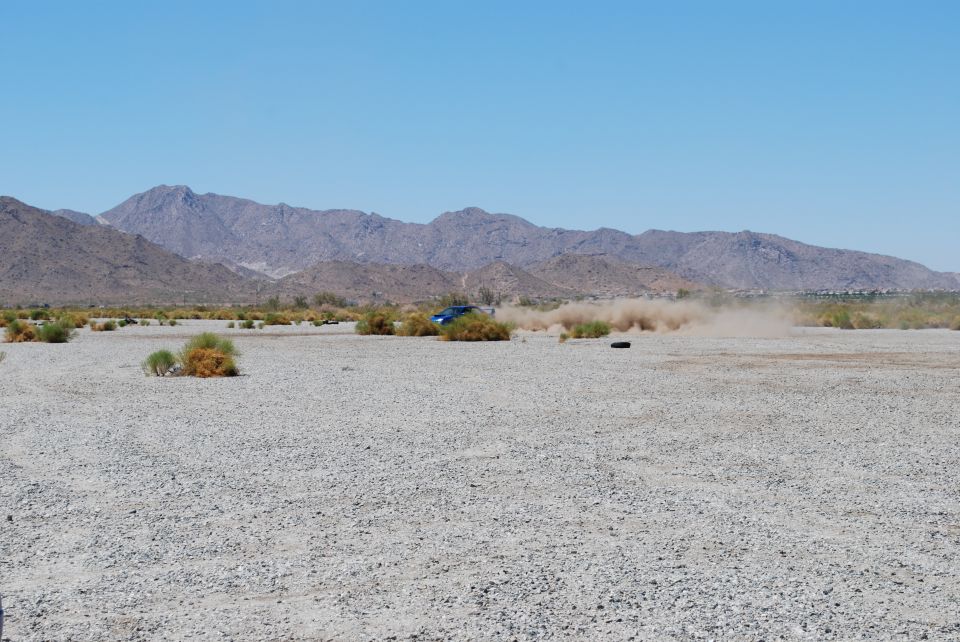 Blue Subaru driving on a gravelly plain, kicking up dust. Mountains in background