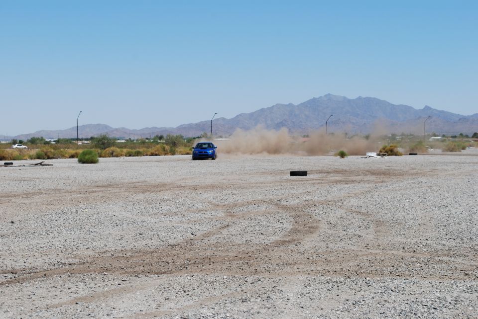 Blue sedan kicking up dust on a gravel lot with mountains in the background