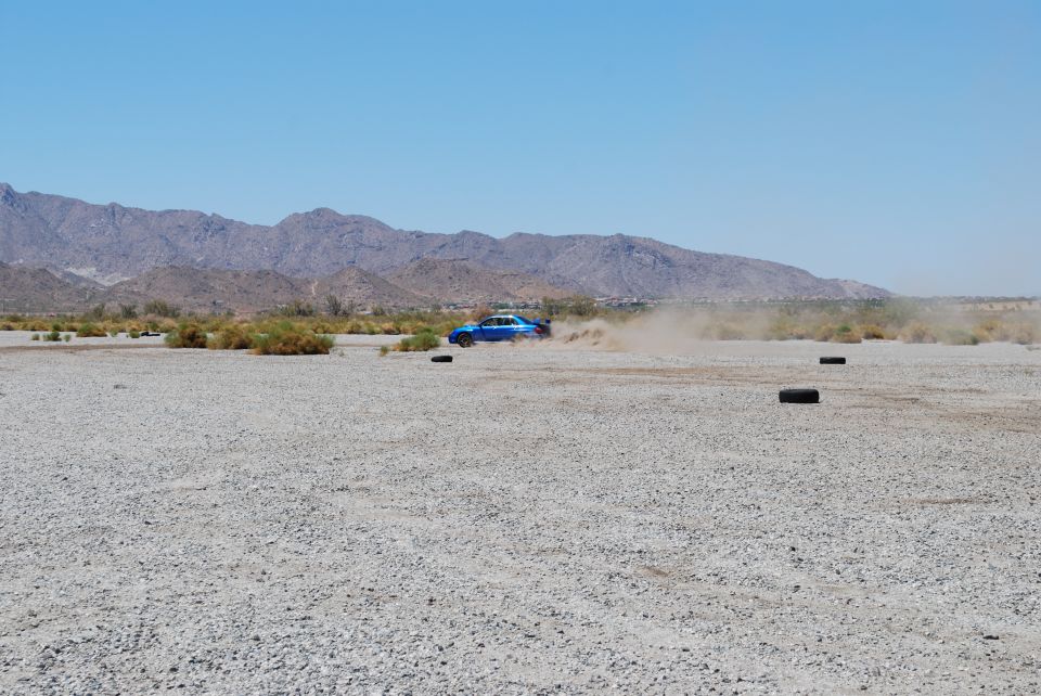 Blue sedan drifting on a dirt track, kicking up dust. Mountains in the background