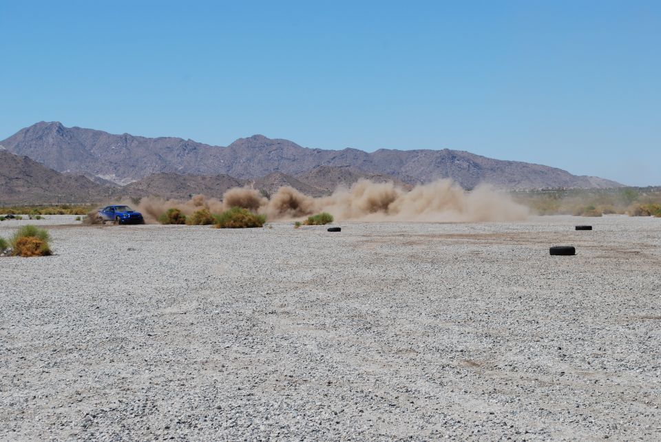 Blue Subaru Impreza driving on a dirt road, kicking up a large dust cloud. Mountains in background