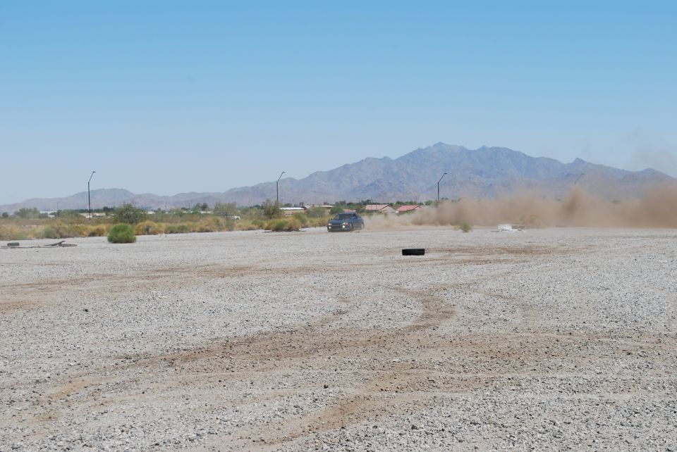 Dark sedan kicking up dust on a gravel lot with mountains in the background