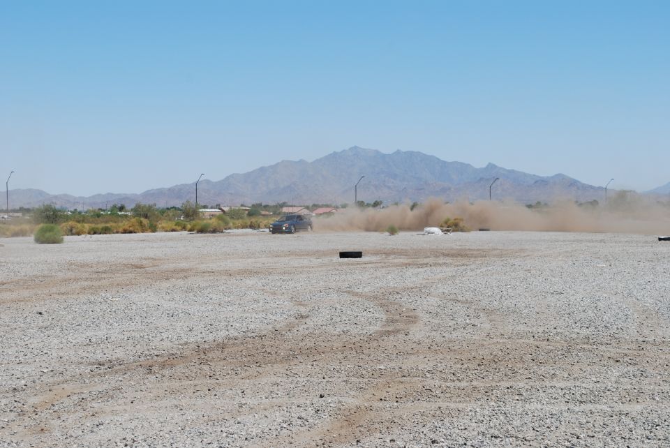 Dark sedan driving on a dusty lot, kicking up a large cloud of dust