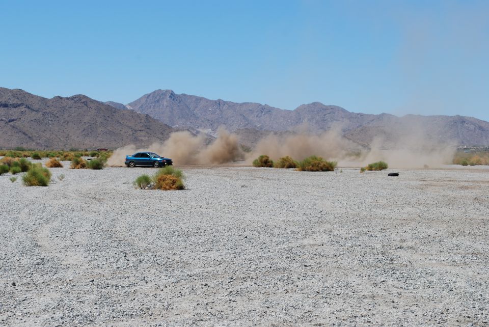 Blue sedan driving on a dusty, gravelly plain with mountains in the background