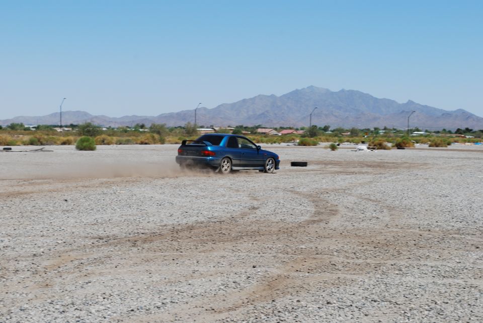 Blue coupe doing a drift in a dusty lot. It has a spoiler and aftermarket wheels