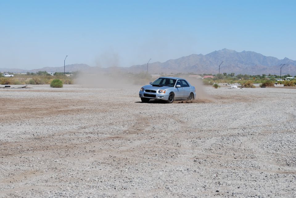 Silver Subaru Impreza drifting on a dirt lot, kicking up dust. Mountains in background