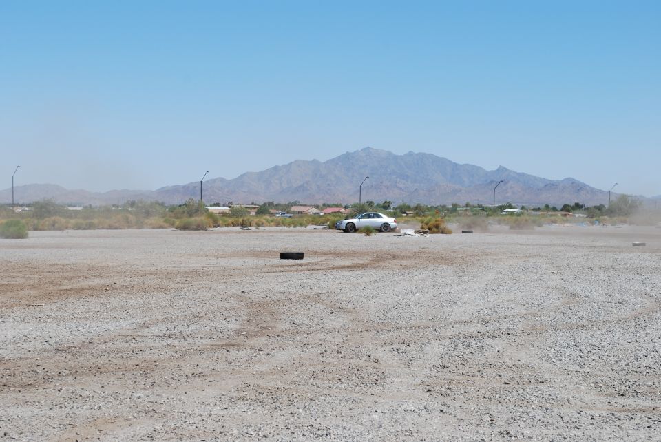 White sedan kicking up dust in a gravel lot with mountains in the background