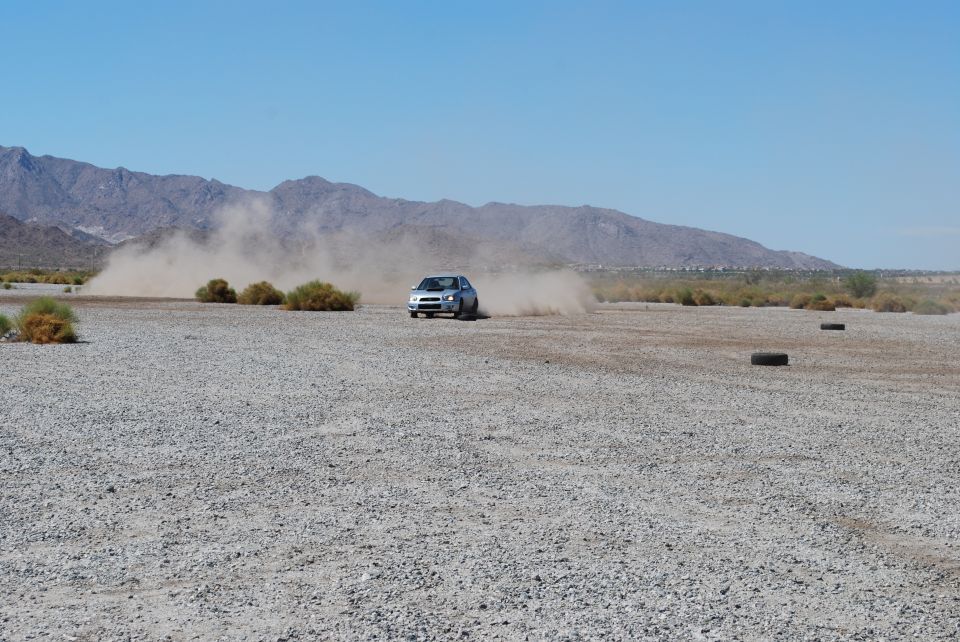 2005 Subaru Impreza drifting on a gravel track, kicking up dust, with mountains in the background