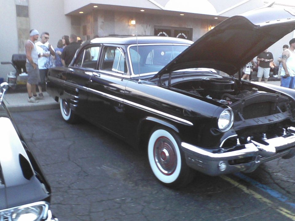 Black 1954 Mercury sedan with hood open, parked outside. Another car is partially visible in the foreground