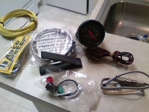 1990 Ford Bronco - An Autometer pyrometer (EGT) gauge kit laid out on a kitchen counter, showing the black-face gauge with red needle and 2-16 scale, its coil…