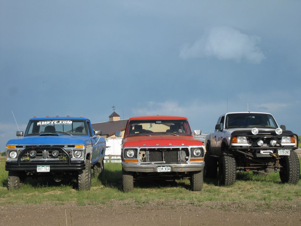 Three trucks parked in a field under a cloudy sky. The leftmost is blue, the middle is red, and the rightmost is silver