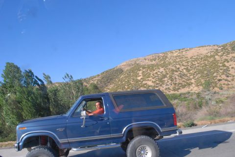 1996 Ford Bronco - A blue SUV with a lifted suspension is driven by a man on a sunny day.