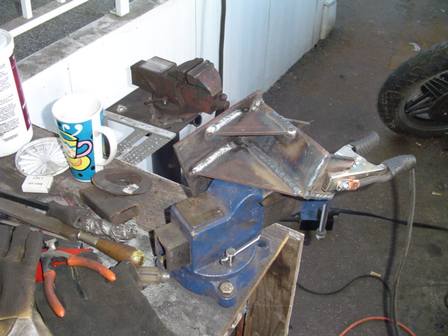 Welded steel bracket clamped in blue bench vise on cluttered workbench