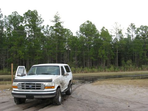 1994 Ford Bronco - A muddy white Ford Bronco is parked on a dirt road with its driver door open.