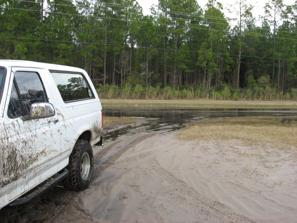 1994 Ford Bronco covered in mud, parked on a dirt track near a wooded area