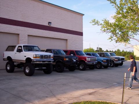 1995 Ford Bronco - Several Ford Broncos and trucks are lined up in a parking lot outside a commercial building.